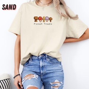 May include: Sand-colored t-shirt with a mushroom graphic and the text "Forest Treats". The graphic features brown, red, and purple mushrooms. The shirt is made of a soft, comfortable material.
