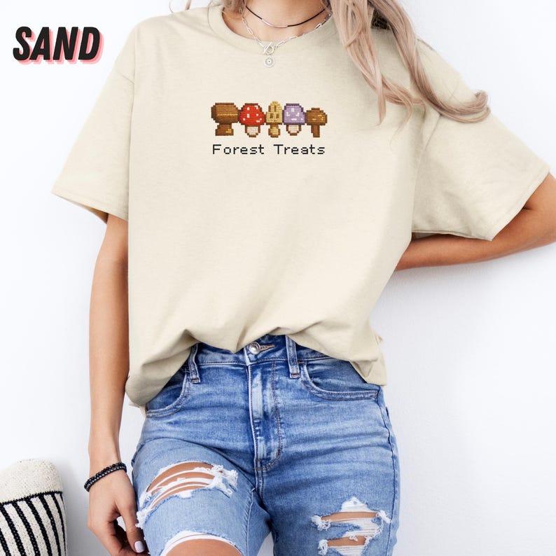 May include: Sand-colored t-shirt with a mushroom graphic and the text "Forest Treats". The graphic features brown, red, and purple mushrooms. The shirt is made of a soft, comfortable material.