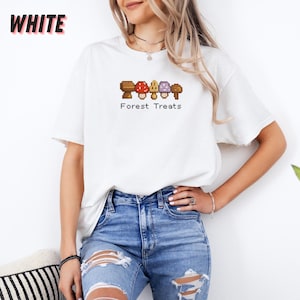 May include: White t-shirt with a pixelated mushroom design and the text "Forest Treats". The mushrooms are brown, red, yellow, and purple. The word "WHITE" is printed in large, bold, black letters in the upper left corner.