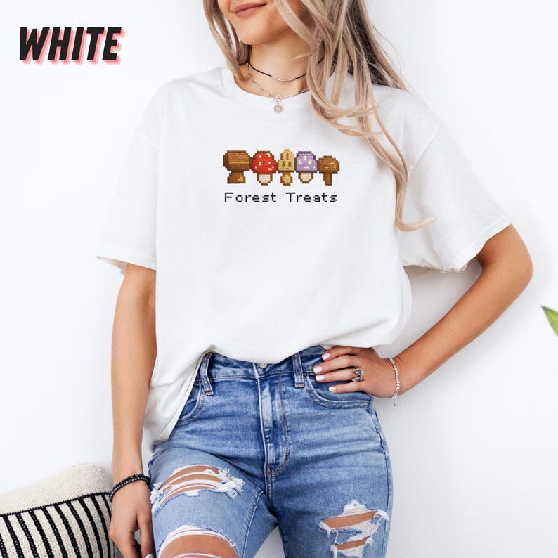 May include: White t-shirt with a pixelated mushroom design and the text "Forest Treats". The mushrooms are brown, red, yellow, and purple. The word "WHITE" is printed in large, bold, black letters in the upper left corner.