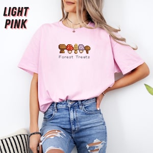May include: Light pink t-shirt with the text "LIGHT PINK" in black block letters. The shirt features a pixelated graphic of mushrooms with the text "Forest Treats" below. The model is wearing blue jeans.