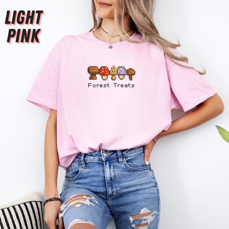 May include: Light pink t-shirt with the text "LIGHT PINK" in black block letters. The shirt features a pixelated graphic of mushrooms with the text "Forest Treats" below. The model is wearing blue jeans.