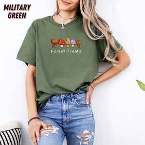 May include: Military green t-shirt with a crew neck and short sleeves. The shirt features a pixelated graphic of five mushrooms in various colors, with the text "Forest Treats" below the design. The shirt is paired with blue jeans.