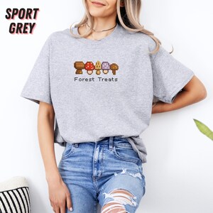 May include: Sport grey t-shirt with a pixelated mushroom design and the text "Forest Treats". The design features brown, red, purple, and tan mushrooms. The shirt is made of a soft material and has short sleeves.