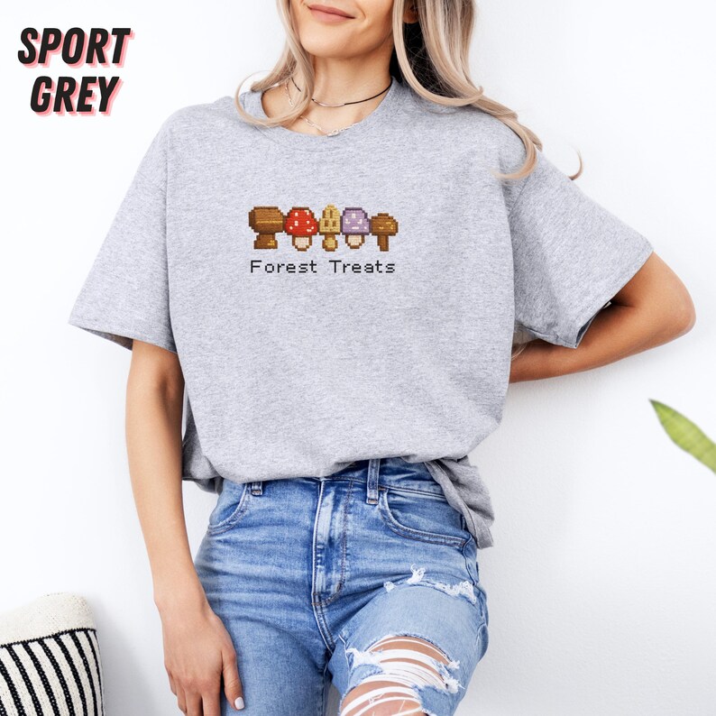 May include: Sport grey t-shirt with a pixelated mushroom design and the text "Forest Treats". The design features brown, red, purple, and tan mushrooms. The shirt is made of a soft material and has short sleeves.