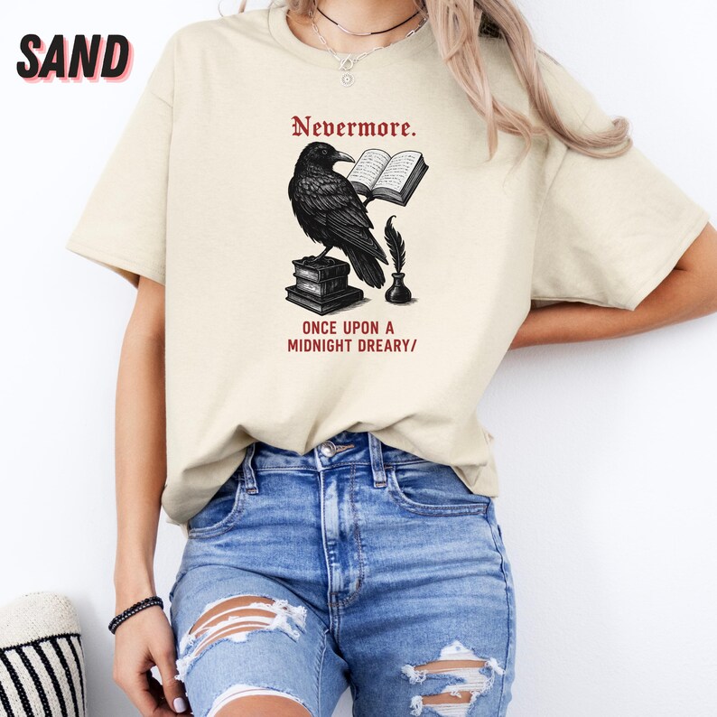 May include: Sand-colored t-shirt featuring a black raven perched on books, with the text "Nevermore." and "ONCE UPON A MIDNIGHT DREARY/". The design includes an open book, quill, and inkwell. The shirt is a casual wear item.