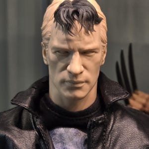 May include: A close-up of a man's face with short, dark hair styled with a side parting. He is wearing a black leather jacket over a blue and white patterned shirt.