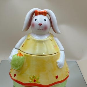 May include: A ceramic bunny cookie jar with a yellow dress, red bow, and green base. The bunny has long white ears, rosy cheeks, and holds a green egg-shaped object. The jar is decorated with floral and heart motifs, perfect for Easter.