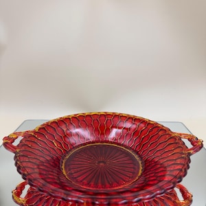 May include: A red glass serving bowl with a honeycomb pattern and gold trim. The bowl has two handles and a circular base. The bowl is sitting on a reflective surface.