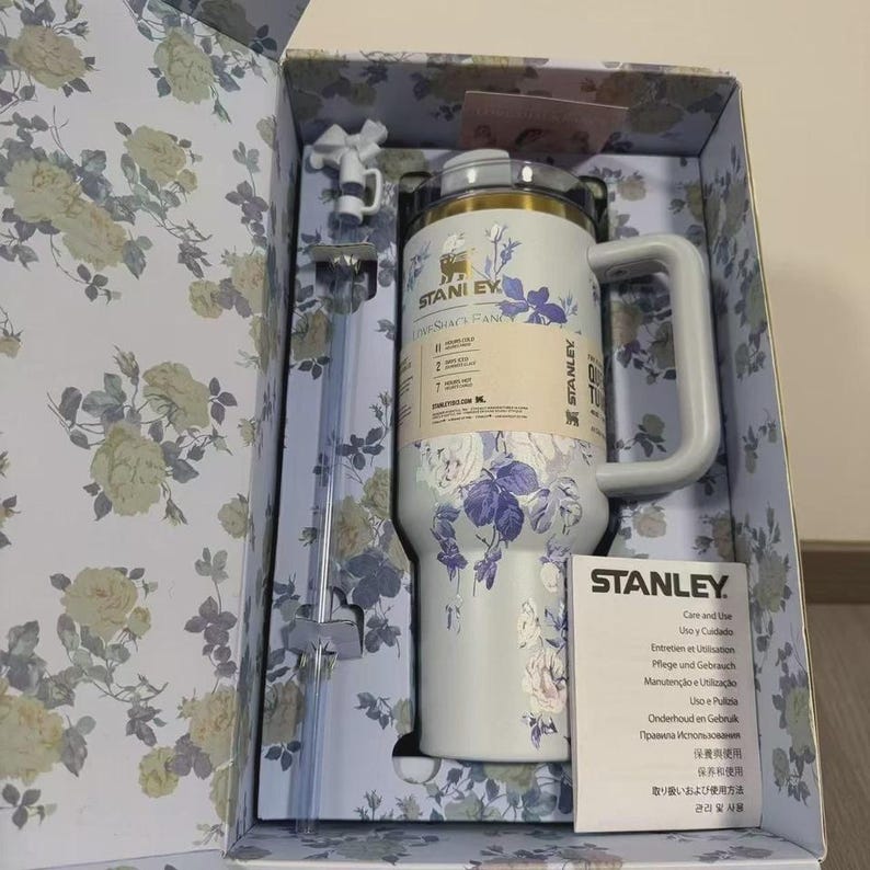 May include: A Stanley insulated mug with a handle, featuring a floral design in white and purple. The mug is in a decorative box with a matching floral pattern. Includes a straw and a small accessory.
