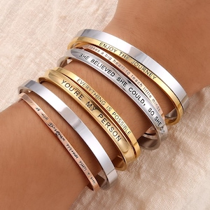 Puede incluir: Un conjunto de cinco pulseras de oro y plata con mensajes inspiradores grabados en ellas. Los mensajes son: "Enjoy the Journey", "She Believed She Could, So She Did", "Everything is Possible", "You're My Person", y "Leave a little sparkle wherever you go".