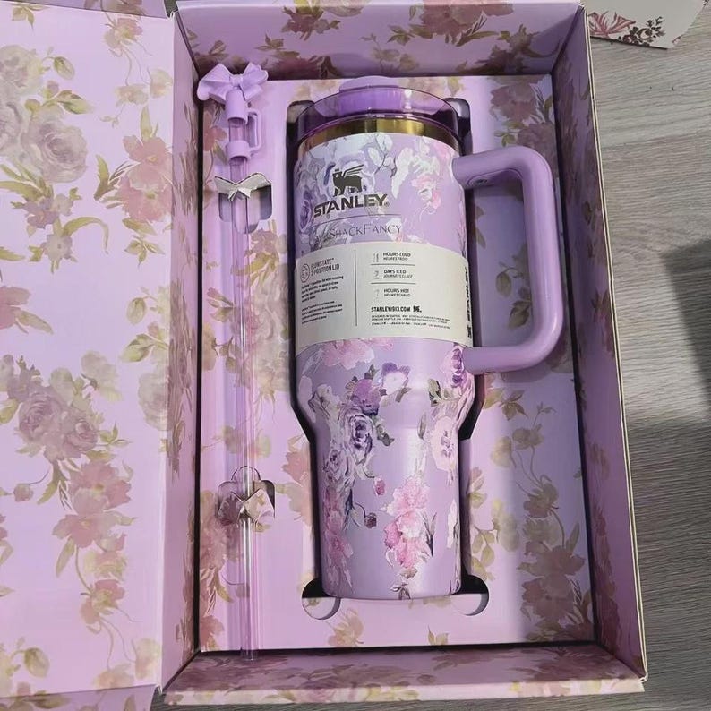 May include: A floral-patterned Stanley Quencher H2.0 FlowState tumbler in a gift box. The tumbler is lavender with a handle and a matching straw. The box and straw have a floral design. The tumbler has the text "STANLEY" and "SHACKFANCY".