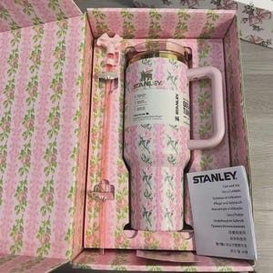 May include: A pink and white floral Stanley tumbler with a handle and straw, presented in a matching floral box. The tumbler has a gold rim and the Stanley logo. The box and tumbler feature a pink floral pattern.