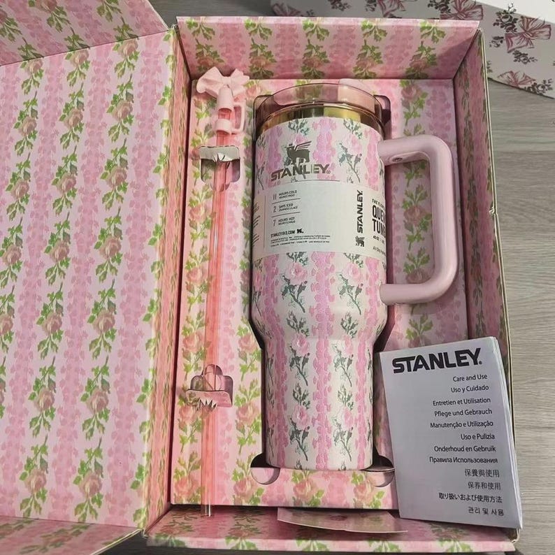 May include: A pink and white floral Stanley tumbler with a handle and straw, presented in a matching floral box. The tumbler has a gold rim and the Stanley logo. The box and tumbler feature a pink floral pattern.