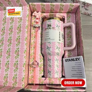 May include: A pink and white floral Stanley tumbler with a handle and a straw, presented in a matching floral box. The tumbler has the Stanley logo and a label. The box has a pink floral pattern. The image also includes a pink straw.
