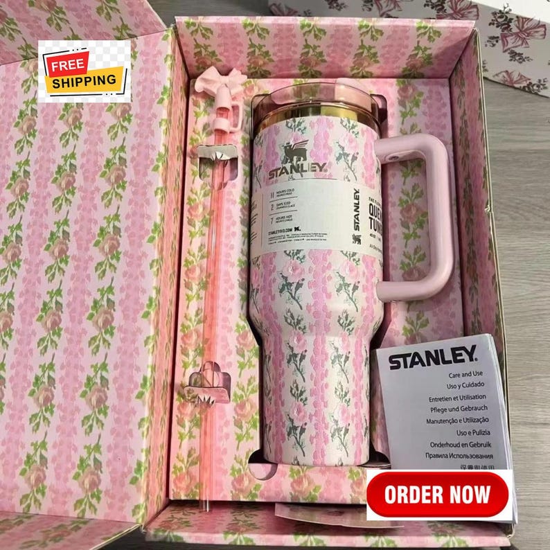 May include: A pink and white floral Stanley tumbler with a handle and a straw, presented in a matching floral box. The tumbler has the Stanley logo and a label. The box has a pink floral pattern. The image also includes a pink straw.
