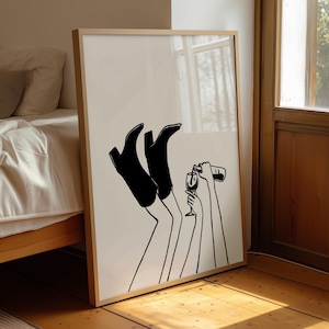 May include: A framed wall art print featuring a minimalist black line drawing on a white background. The illustration depicts a pair of legs wearing black boots pointed upward, alongside two arms, one holding a wine bottle and one holding a glass.