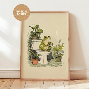 Op de afbeelding: Een ingelijste kunstdruk met een groene kikker die op een toilet zit en een kopje vasthoudt. De illustratie bevat potplanten en een neutraal kleurenpalet. De print is gelabeld met "PHYSICAL POSTER" in een cirkelvormig ontwerp.