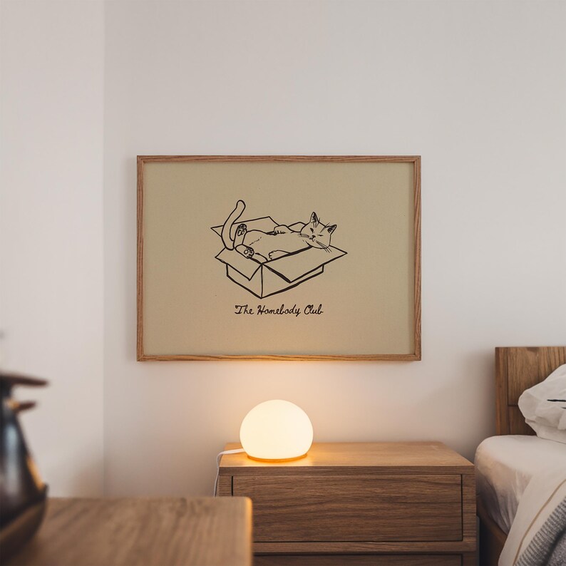 Minimal black line drawing of a cat lounging in a box, styled as a funny cat poster with homebody decor charm for a cozy book nook poster space, text reads “The Homebody Club”.