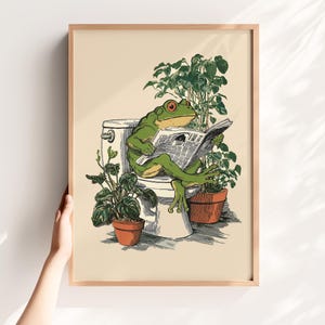 Könnte beinhalten: Gerahmter Kunstdruck mit einer grünen Froschfigur mit roten Augen, die auf einer Toilette sitzt und Zeitung liest. Topfpflanzen umgeben die Toilette. Das Kunstwerk hat einen skurrilen, cartoonartigen Stil mit hellem Beige-Hintergrund.