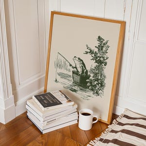 Hand drawn poster of a fishing frog sitting in a bucket by the water, ink drawing with vintage line art style, frog wall art for lake house decor, cabin decor, and fishing wall art.
