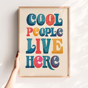 May include: A framed print featuring the phrase "COOL PEOPLE LIVE HERE" in a retro font. The text is in vibrant colours including blue, red, orange, and pink, set against a cream backdrop, within a light brown wooden frame.