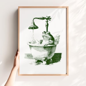 Peut inclure: Impression encadrée en vert et blanc d'une grenouille dans une baignoire. La grenouille est représentée dans un style vintage, prenant un bain sous une pomme de douche. L'œuvre est de style botanique, avec un cadre en bois clair.
