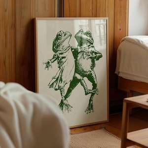 May include: A framed wall art print featuring a dark green ink illustration of two frogs standing upright. One frog wears a cape and the other wears a bow tie. The frogs are depicted in a dancing pose against a light cream-colored background.