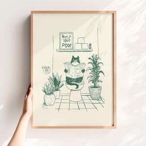 Op de afbeelding: Een ingelijste print met een groene inktillustratie van een zwarte kat die een krant leest op een toilet. Het kunstwerk bevat de tekst "Have a Nice Poop". De badkamer scène bevat ook potplanten en andere armaturen, allemaal in een speelse stijl.