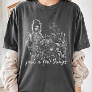 Puede incluir: Una camiseta gris carbón con una ilustración gráfica de una persona sosteniendo una bebida junto a un carrito de compras lleno de artículos de Halloween, incluyendo calabazas, fantasmas y murciélagos. El texto "just a few things" está impreso en blanco.