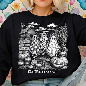 Puede incluir: Una sudadera negra con un estampado gráfico blanco de tres fantasmas frente a un granero y un campo de maíz. Los fantasmas tienen patrones de cuadros, leopardo y lunares. El texto dice PUMPKINS, HAY RIDES, CORN MAZE y tis the season.