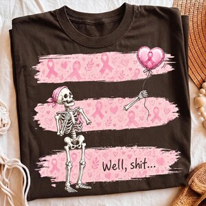 Puede incluir: Una camiseta marrón oscuro con un gráfico de un esqueleto que lleva un pañuelo rosa. El esqueleto sostiene un globo rosa en forma de corazón con una cinta. Tres bandas horizontales de pinceladas rosas contienen patrones de corazones y lazos de cáncer de mama. El texto "Well, shit..." está impreso en negro.