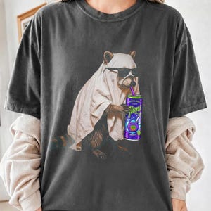 Op de afbeelding: Een antracietgrijs t-shirt met een afbeelding van een wasbeer die een wit laken als spookkostuum en een zwarte zonnebril draagt. De wasbeer houdt een paars energiedrankblikje met een rietje vast, waarop de tekst Glani en Witch's Brew staat.