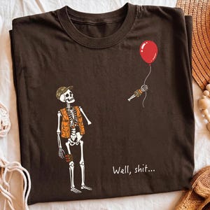 May include: A dark brown t-shirt featuring a graphic of a skeleton wearing a camouflage cap and an orange hunting vest. The skeleton is looking up at a floating red balloon held by a detached skeletal hand. The text "Well, shit..." is printed below.