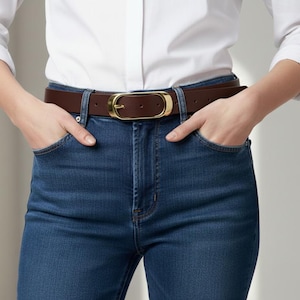May include: A brown leather belt with a gold-colored oval buckle is worn with blue jeans and a white button-down shirt. The belt is approximately 1.5 inches wide. The person's hands are in the pockets of the jeans.