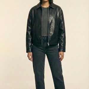 May include: A woman wearing a black leather jacket, a gray t-shirt, and dark gray jeans. The jacket has a collar and a zipper closure. The jeans are straight-legged, and the boots are black. The brand name "VELVISTACO" is visible in the top left corner.