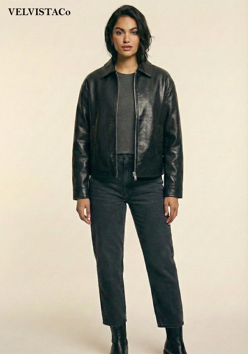 May include: A woman wearing a black leather jacket, a gray t-shirt, and dark gray jeans. The jacket has a collar and a zipper closure. The jeans are straight-legged, and the boots are black. The brand name "VELVISTACO" is visible in the top left corner.
