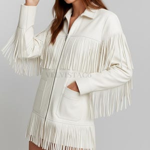 May include: A white jacket with a full-length front zipper, a classic collar, and two front pockets. The jacket features long fringe detailing across the chest, sleeves, and hemline. The text VELVISTACO is printed in a light gray font on the front.