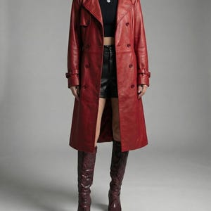 Puede incluir: Un abrigo largo de cuero rojo con diseño cruzado. El abrigo presenta un cuello con solapas, cierres de botones y detalles de cinturón en los puños. La modelo lleva shorts negros y botas altas burdeos. La marca "VELVISTACO" es visible en la esquina superior izquierda.