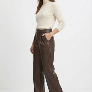 May include: A woman wearing a cream-colored turtleneck sweater and brown leather pants. The pants are high-waisted with a wide leg. The outfit is simple and elegant, suitable for various occasions. The brand name "VELVISTACO" is visible in the top left corner.