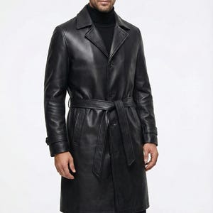 Könnte beinhalten: Schwarzer Ledertrenchcoat mit Gürtel, Knopfverschluss und Revers. Der Mantel hat lange Ärmel und ein klassisches Design. Der Markenname "VELVISTACO" ist oben links sichtbar.