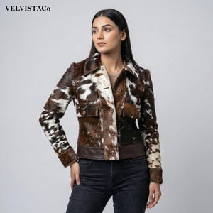 May include: A brown and white cowhide jacket with a button-up front, two chest pockets, and a brown leather trim. The jacket has a collared neckline and long sleeves. The brand name "VELVISTACO" is in the top left corner.
