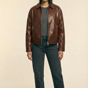May include: A brown leather jacket with a zipper closure and a collared neckline. The jacket is paired with a gray t-shirt and dark gray jeans. The brand name "VELVISTACO" is visible in the top left corner.