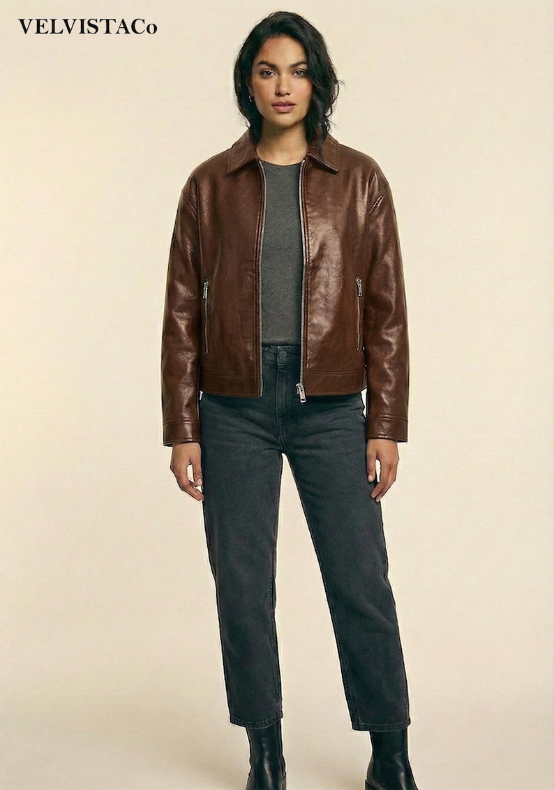 May include: A brown leather jacket with a zipper closure and a collared neckline. The jacket is paired with a gray t-shirt and dark gray jeans. The brand name "VELVISTACO" is visible in the top left corner.