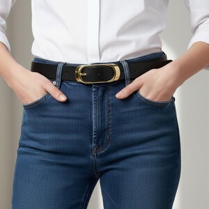 May include: A black leather belt with a gold-colored buckle is worn with blue jeans and a white button-down shirt. The buckle is oval-shaped. The person's hands are in the pockets of the jeans.