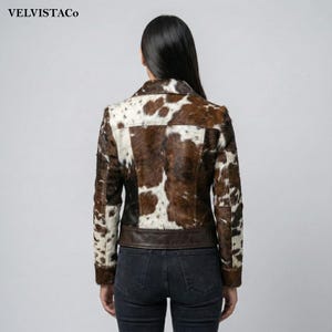 May include: A brown and white cowhide jacket with a dark brown leather trim at the waist. The jacket has a classic collar and button closure. The brand name "VELVISTACO" is visible in the top left corner.