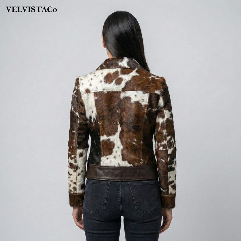 May include: A brown and white cowhide jacket with a dark brown leather trim at the waist. The jacket has a classic collar and button closure. The brand name "VELVISTACO" is visible in the top left corner.