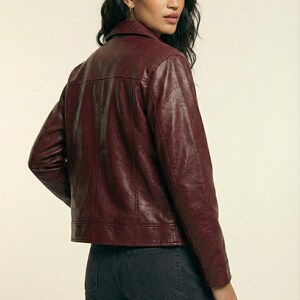 May include: A burgundy leather jacket with a collared neckline and long sleeves. The jacket has a classic design with a button closure and a tailored fit. The model is wearing dark wash jeans. The brand name "VELVISTACO" is visible in the top left corner.