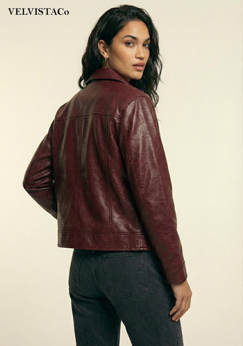 May include: A burgundy leather jacket with a collared neckline and long sleeves. The jacket has a classic design with a button closure and a tailored fit. The model is wearing dark wash jeans. The brand name "VELVISTACO" is visible in the top left corner.