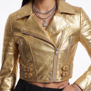 May include: A cropped gold metallic biker jacket with a notched collar, asymmetrical zipper, and quilted shoulder detailing. The jacket features silver hardware, including zippers, snaps, and buckles. The brand name "VELVISTACO" is visible at the top.